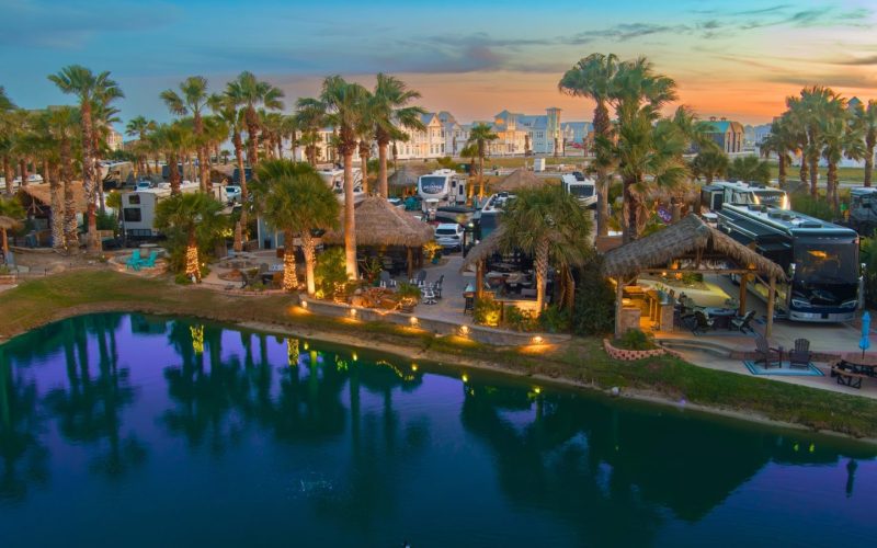 Beachfront RV resort with palm trees and water at sunset.