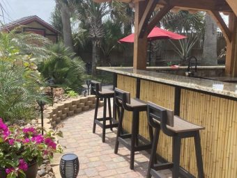 Outdoor bar area with bamboo counter, high stools, surrounded by tropical plants and flowers, featuring patio umbrellas, ideal for relaxation at Gulf Waters Beach Front RV Resort.