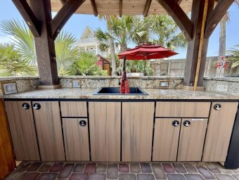 Outdoor kitchen with granite countertop, sink, and storage cabinets, surrounded by palm trees and a red umbrella, showcasing amenities at Gulf Waters Beach Front RV Resort.